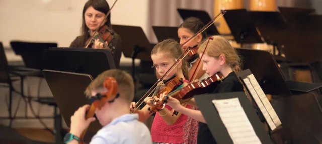 Viele Kinder spielen verschiedene Streichinstrumente vor einer Dirigentin (Musikschullehrerin) - Streicherspielkreis