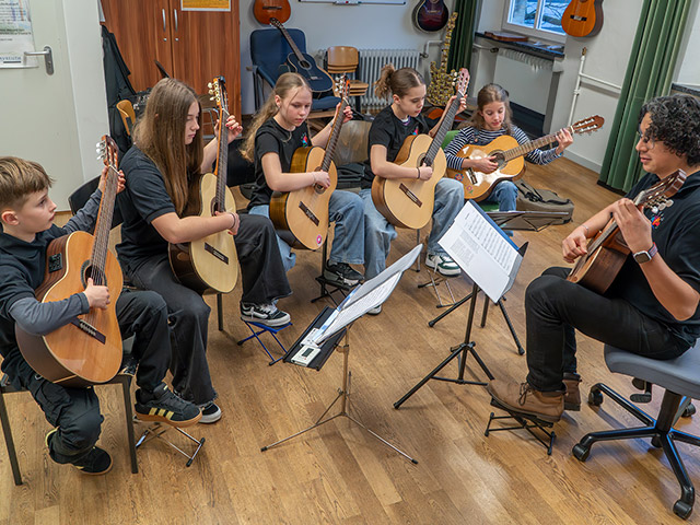 Fünf Kinder sitzen vor Musiklehrer mit Gitarren und spielen zusammen - Gitarrenorchester