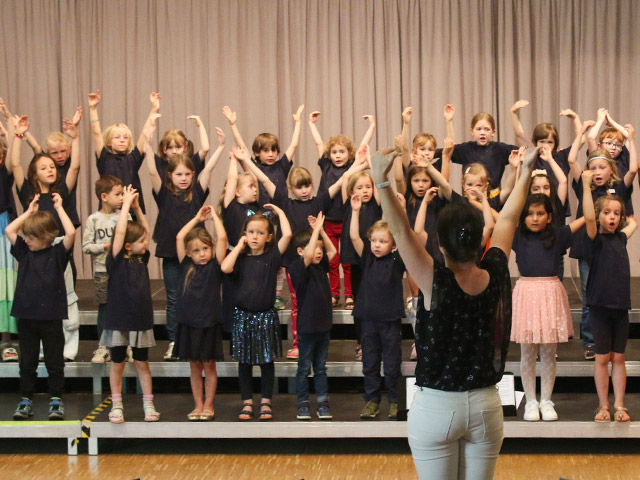 Kinderchor 1 & 2: Kinder auf Bühne mit Musiklehrerin