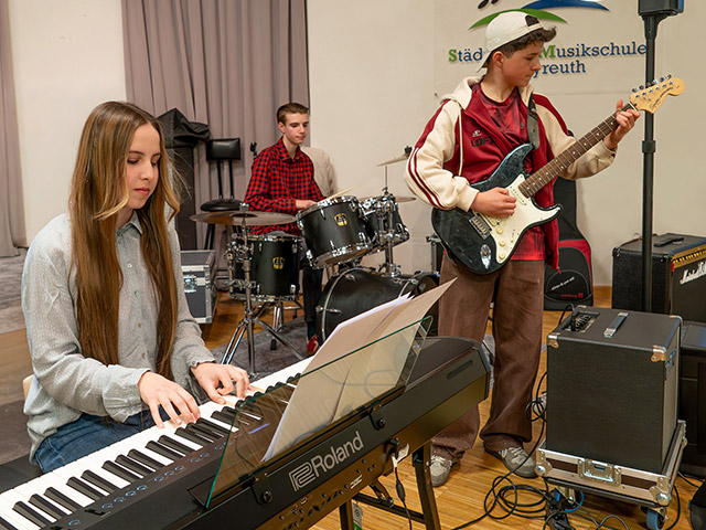 Ein Mädchen spielt Keyboard, ein Junge mit Cap spielt E-Gitarre und im Hintergrund spielt ein weiterer Junge Schlagzeug - Musikschulband