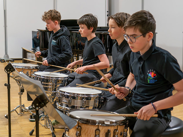 Schwarz gekleidete Jungs spielen sitzend vor Trommeln - Percussionensemble