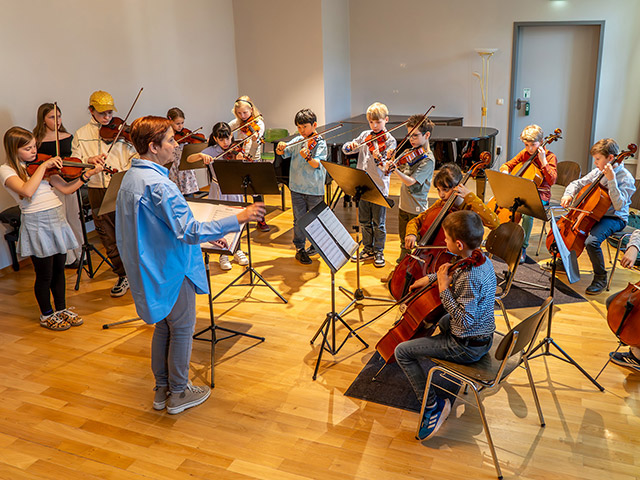 Viele Kinder spielen verschiedene Streichinstrumente vor einer Dirigentin (Musikschullehrerin) - Streicherspielkreis